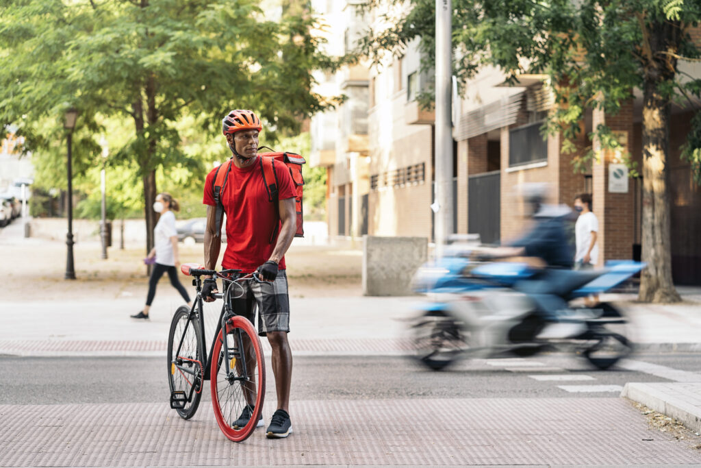 Adult delivery man carrying a package and using a safety helmet crossing the city with his bike.