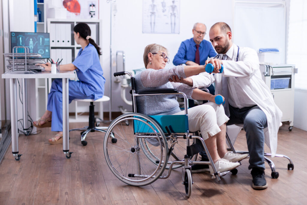 Disabled woman doing recovery with dumbbells and help from doctor in hospital. Man with disabilities ,walking frame sitting in hospital bed. Health care system, patients.