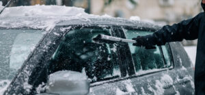 Man hand cleaning car window from snow and ice with brush and scraper tool while it's snowing.